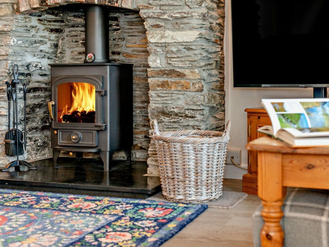 A stone wall, a black wood-burning stove with a visible fire, a wicker basket, a wooden table with a book, and a television.