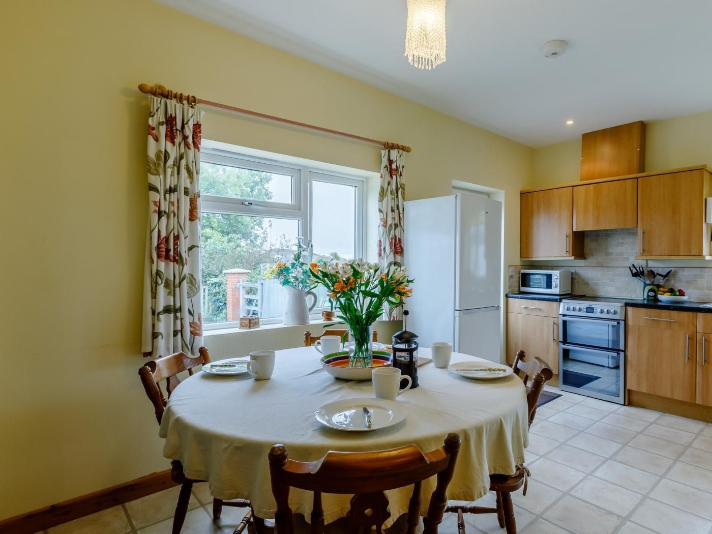 A dining area with a round table, chairs, a vase of flowers, and a window with curtains.