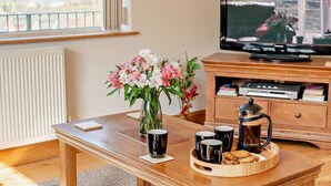 Ein Holztisch mit einer Kaffeekanne, Tassen, Keksen und einer Blumenvase. An der Wand befindet sich ein Fernseher.