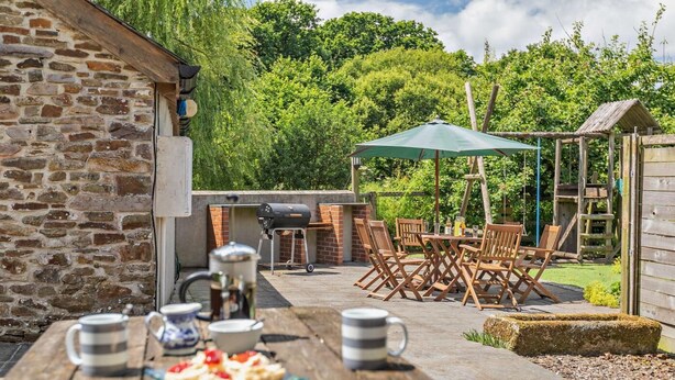 A stone building with a barbecue area, outdoor seating, and a table with food and drinks. - 3 Bed in Winkleigh (oc-1450b) (Iddesleigh)