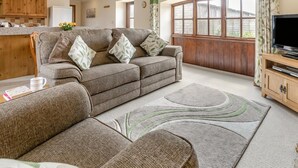 A living room with a sofa, a television, a wooden cabinet, and a window with curtains. - 1 Bed in Helston (oc-bbird) (Saint Martin)