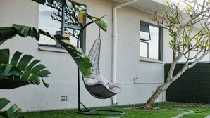 A hanging chair in a garden area with a white building, a tree, and a well-maintained lawn. - The Valley Escape - Unit 2 (Gqeberha)
