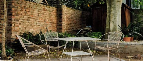 A metal table and chairs set on a paved area with a brick wall and greenery in the background.
