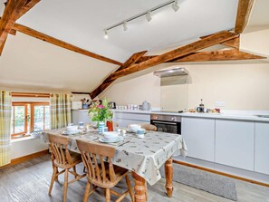 Comedor con una mesa dispuesta para cuatro personas, sillas de madera, cocina con armarios blancos y una ventana con cortinas.