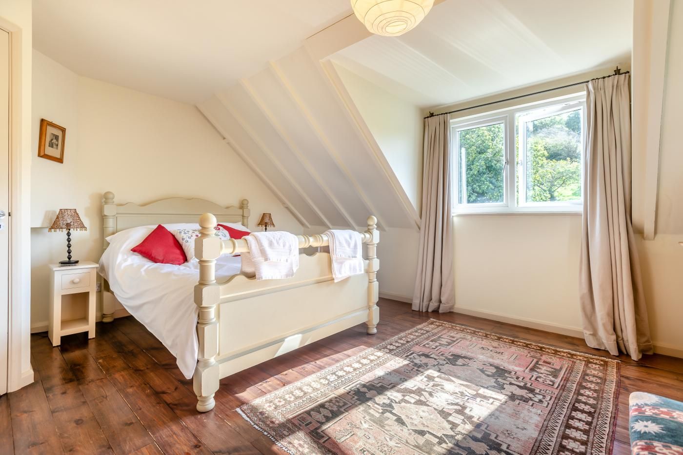 Ein Schlafzimmer mit schrägverdeckter Decke, einem Bett mit weißer Bettwäsche und roten Kissen, einem Nachttisch mit einer Lampe und einem Fenster mit Vorhängen.