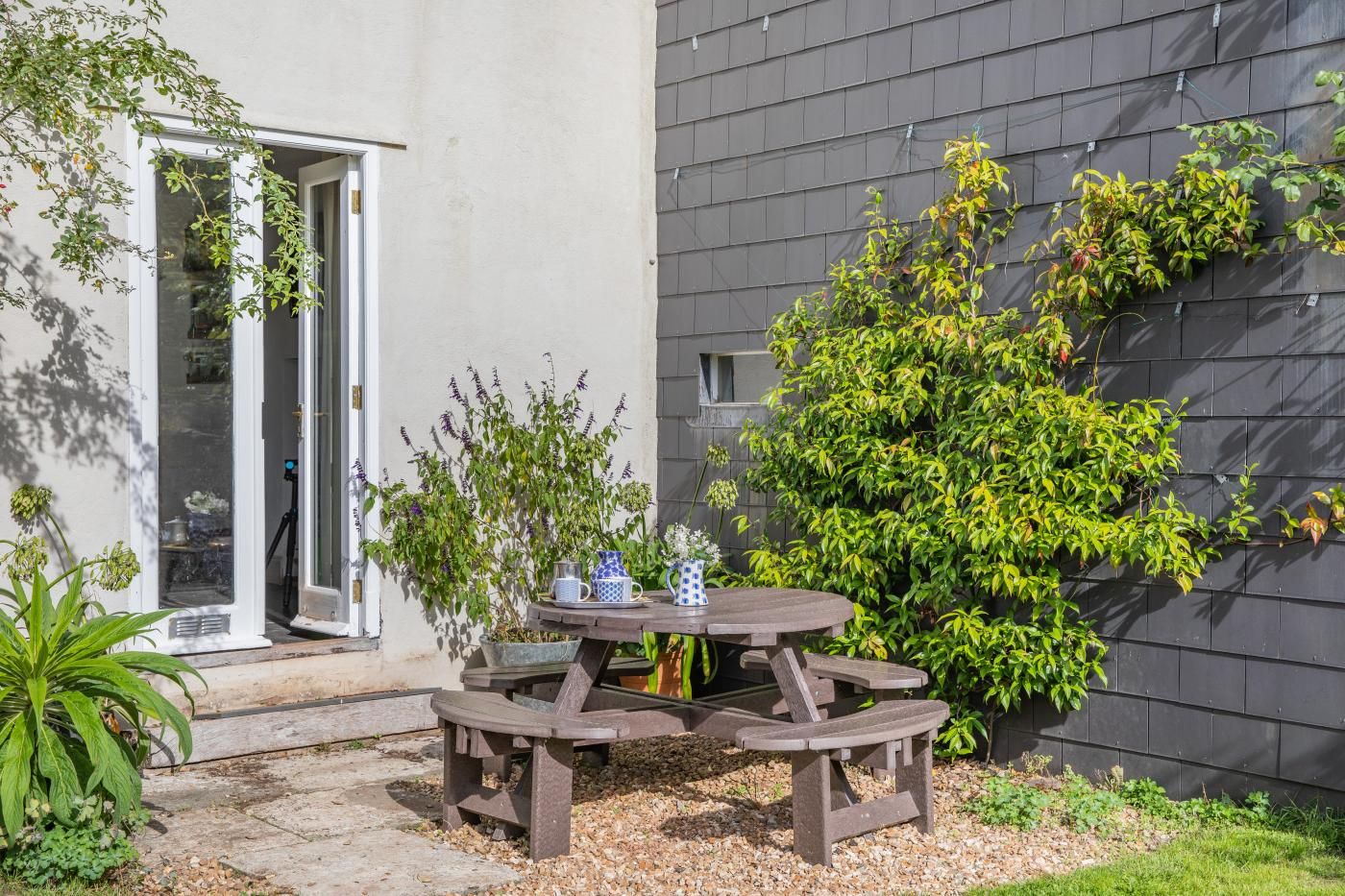 An outdoor patio with a wooden table and benches, surrounded by potted plants and a small tree.