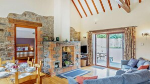A cozy living room with a stone fireplace, a dining area with a wooden table and chairs, a blue sofa, and a large sliding glass door leading to an outdoor patio. - 3 Bed in Bideford (oc-burfl) (Higher Clovelly)