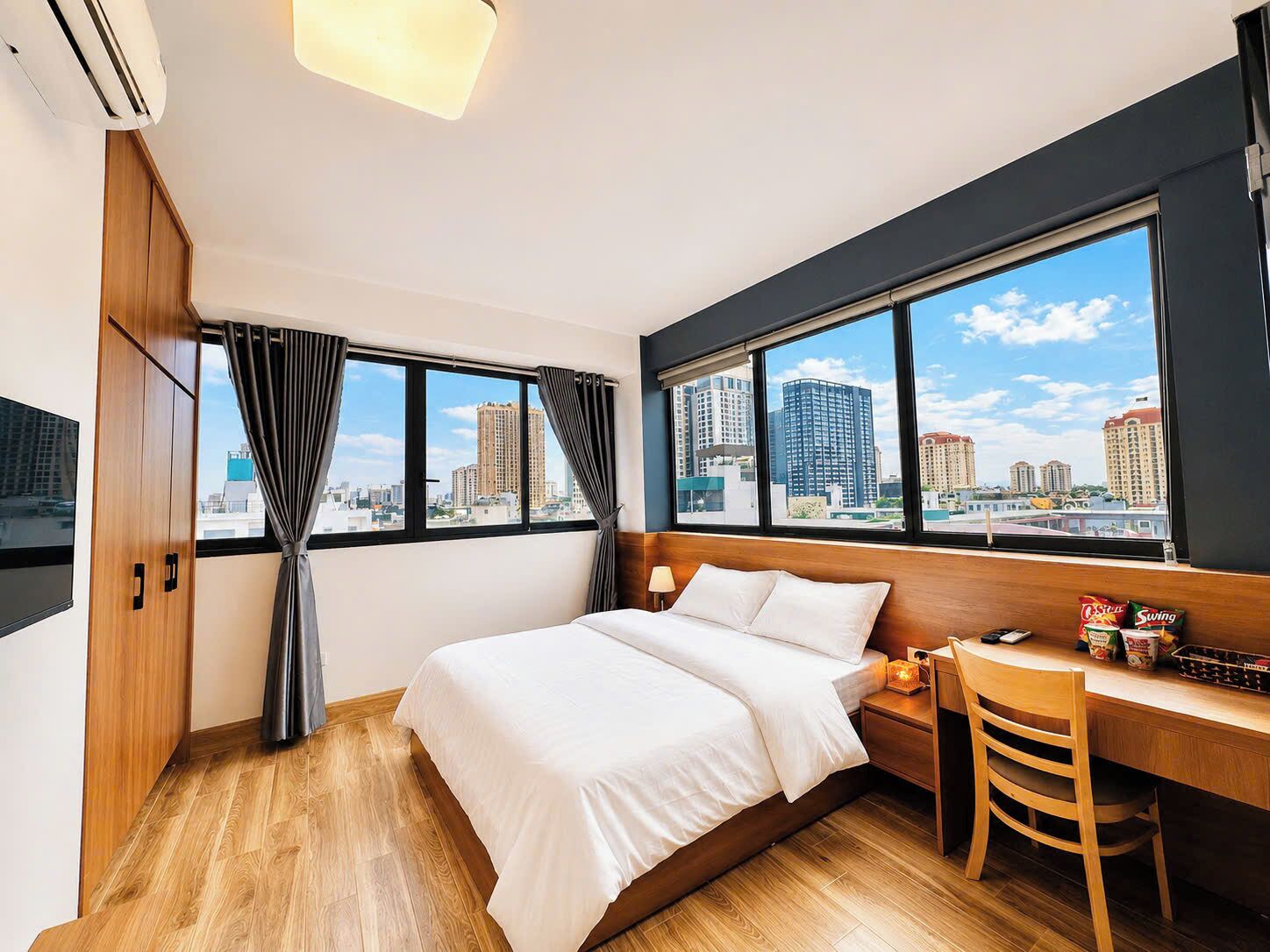 A hotel room with a bed, a desk, a chair, a TV, and large windows with a city view.