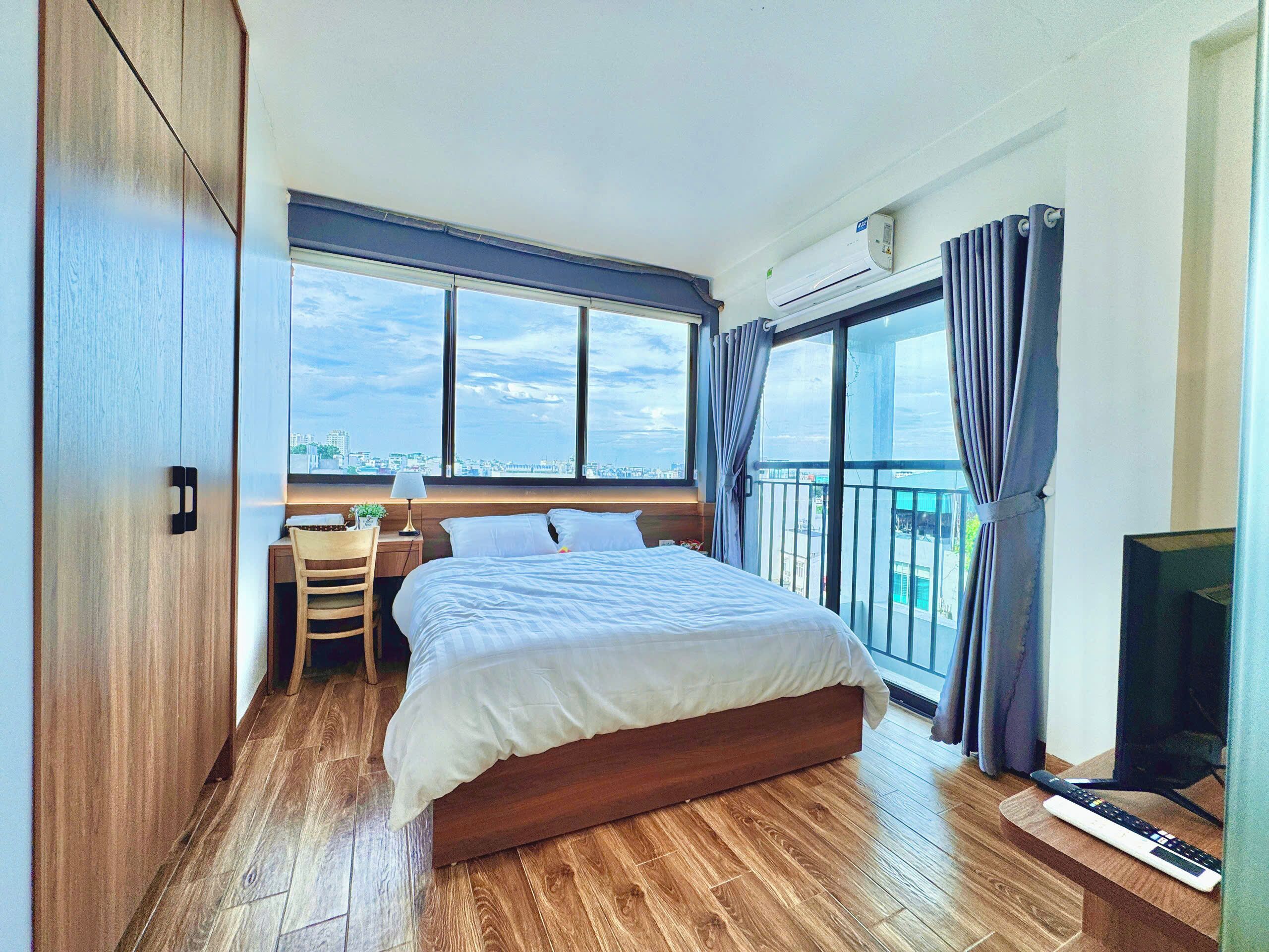 A bedroom with a bed, a desk, a chair, a TV, and a window with a city view.