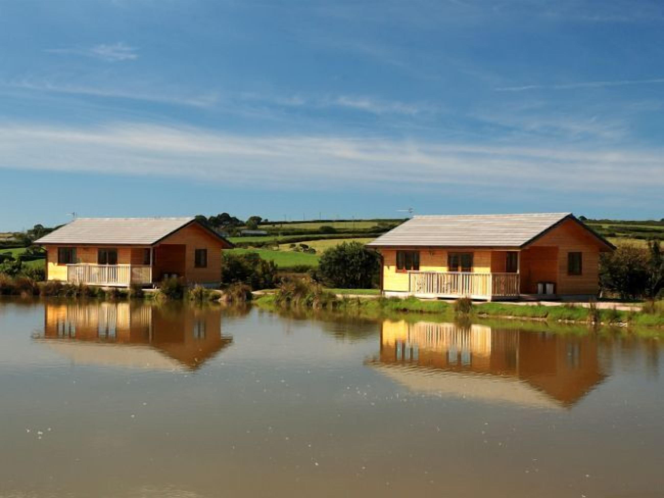 Deux cabanes en bois dotées de balcons se reflètent dans l’eau calme d’un plan d’eau, le tout étant entouré de verdure.