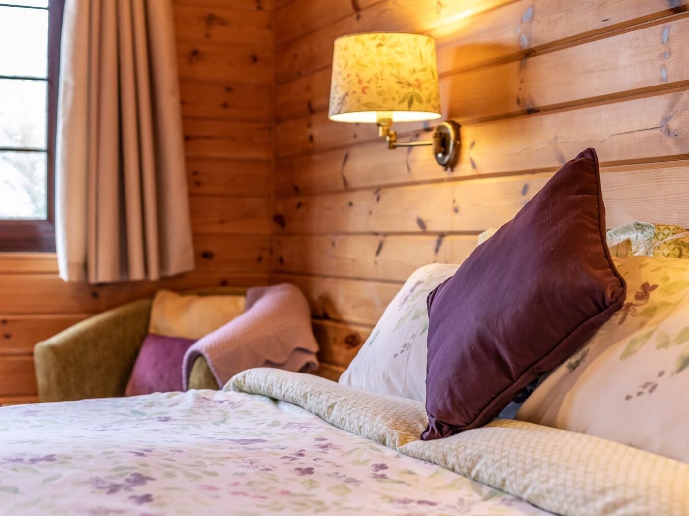 A wooden cabin room with a bed, pillows, a lamp, and a window with curtains.