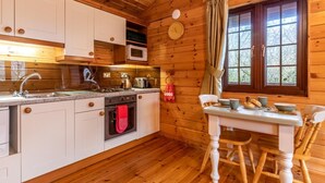 A wooden cabin interior with a kitchen, dining table, chairs, and a window with curtains. - 1 Bed in Bideford (oc-00355) (Higher Clovelly)