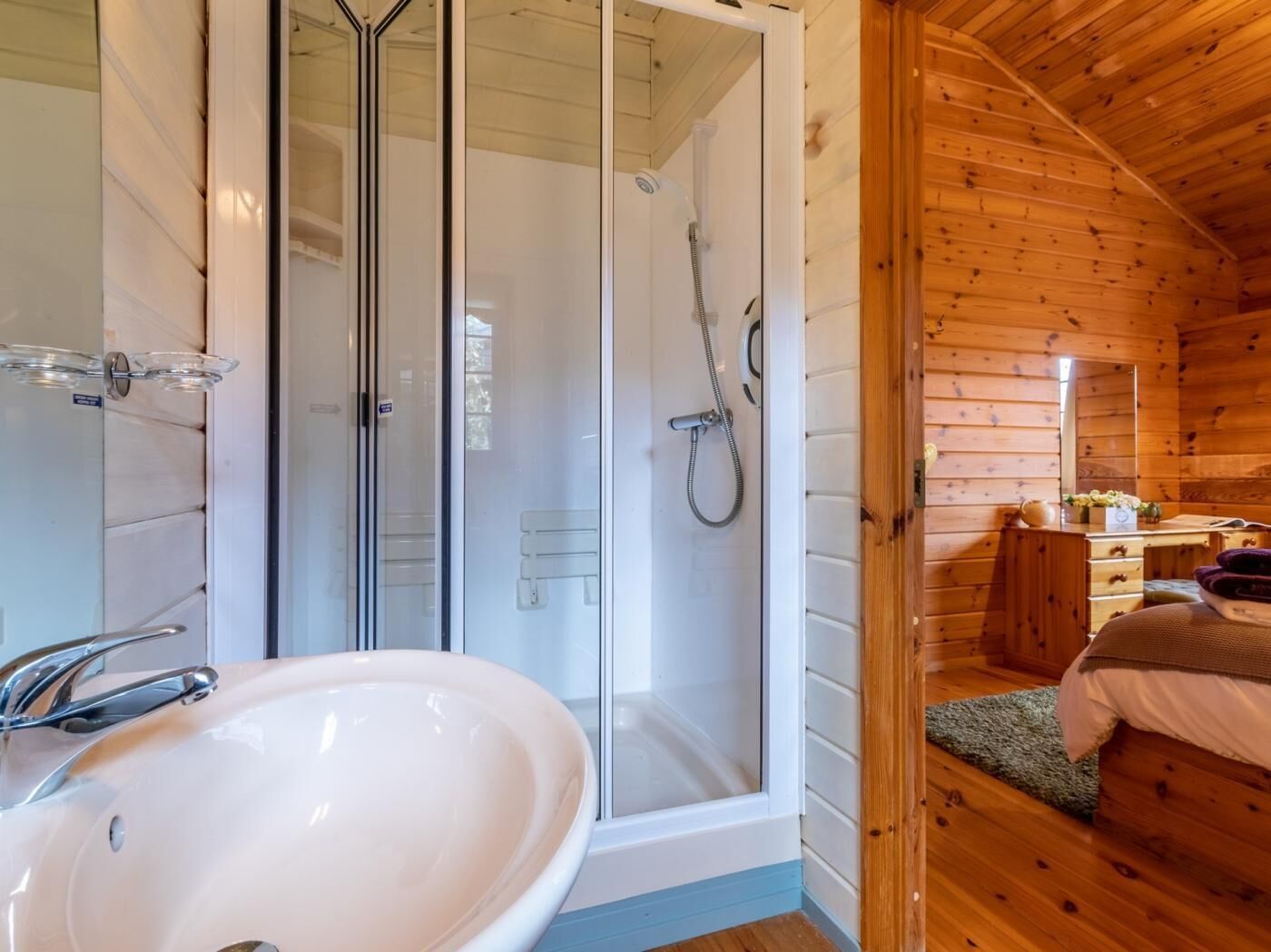 A wooden cabin interior with a glass-enclosed shower, a white sink, and a wooden bed.