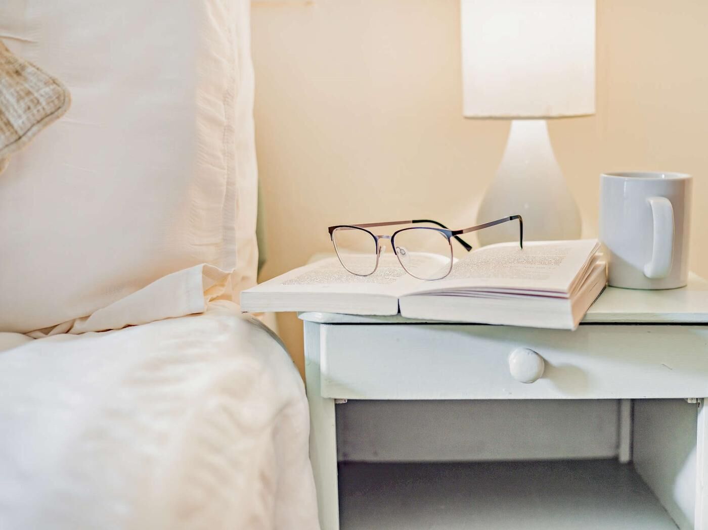 Un lit bien fait, avec du linge de lit blanc, une table de chevet sur laquelle se trouvent un livre, des lunettes et une tasse blanche.