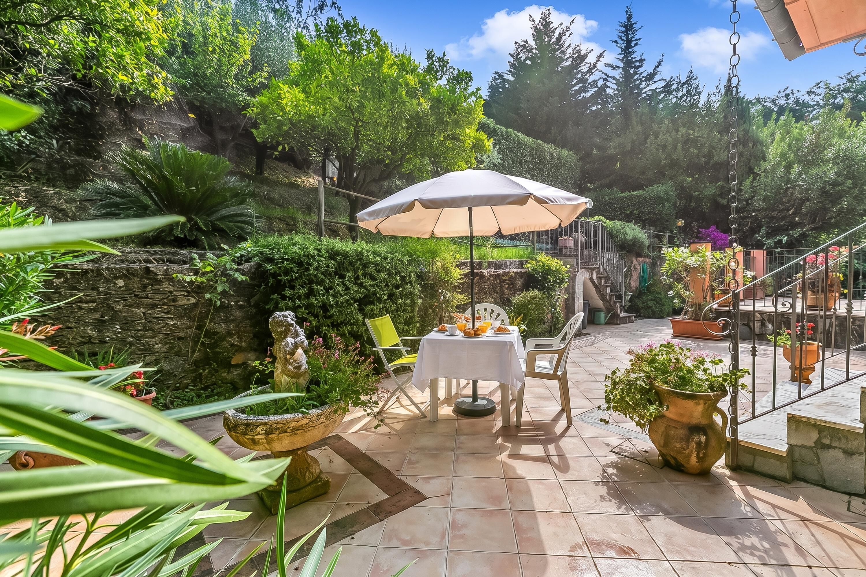 Patio esterno con tavolo e sedie sotto una tettoia, circondato da piante in vaso e un muro in pietra.