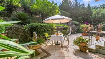 Patio esterno con tavolo e sedie sotto una tettoia, circondato da piante in vaso e un muro in pietra.