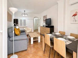 A modern living room with a grey sofa, a white armchair, a wooden coffee table, and a dining area with a dark wooden table set for four. - Las Dunas, T1 (Punta del Moral)