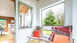 Een kamer met twee stoelen met houten frames, uitzicht op een bos door een groot raam, en een deur naar een andere kamer.