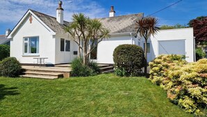 Une maison de plain-pied à l’extérieur blanc, dotée d’une cheminée, d’un petit porche et d’un jardin bien entretenu avec divers arbustes et un palmier.