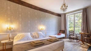 Ein Schlafzimmer mit einem großen Bett, zwei Nachttischen, einer Kandelaberleuchte, einem Fenster mit Vorhängen und Blick auf Grünfläche.