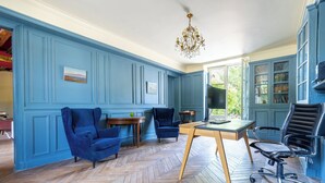 Ein Zimmer mit blauen Wandverkleidungen, einem Holzboden im Fischgrätmuster, einer Chandelüster und einem Schreibtisch mit Stuhl.