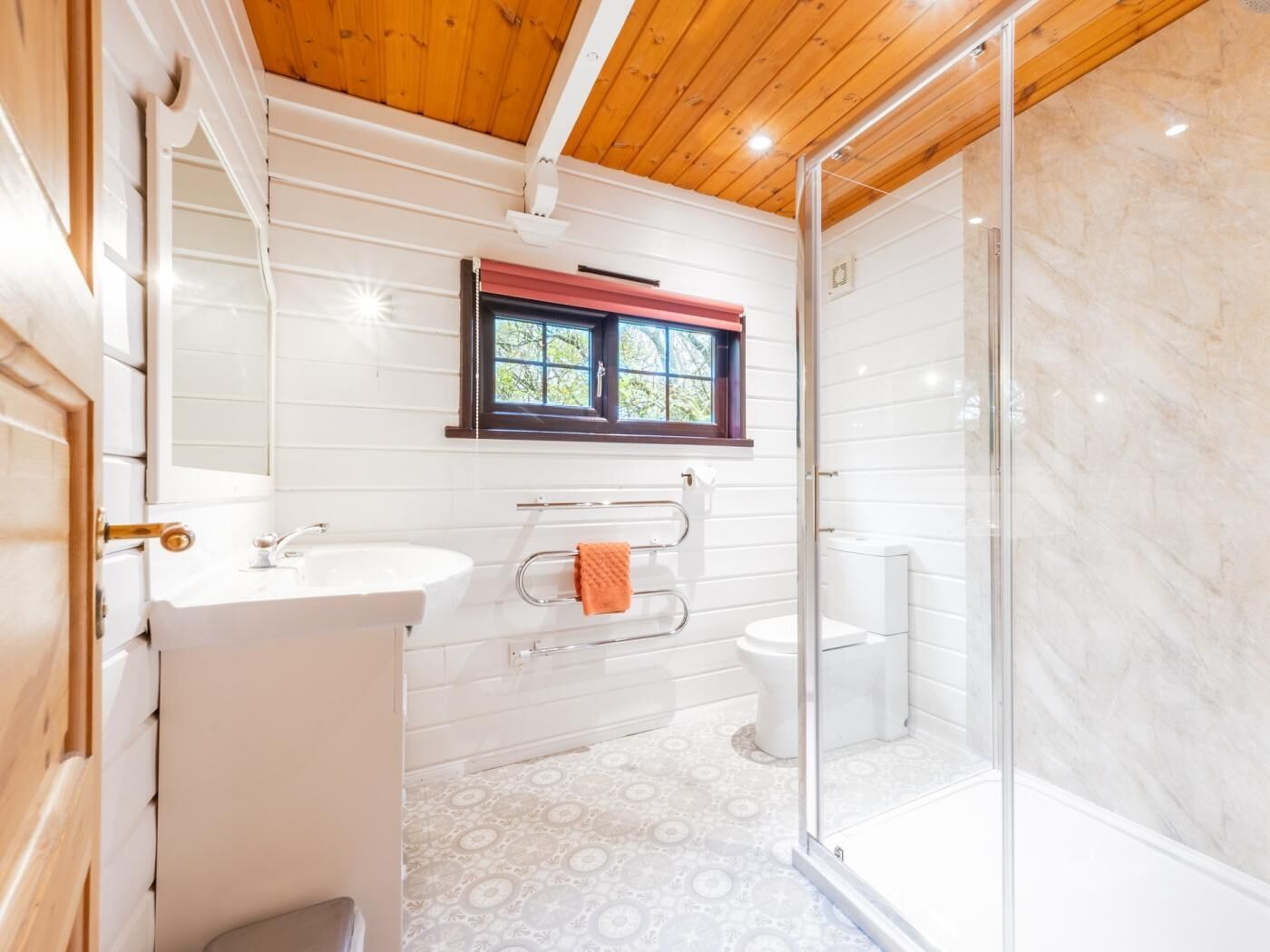 Een badkamer met een witte wasbak, toilet en douchegedeelte, houten plafond en vloertegels met een patroon.