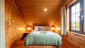 A wooden cabin bedroom with a bed, two bedside lamps, a window, and a blue rug. - 3 Bed in Bideford (oc-00369) (Higher Clovelly)