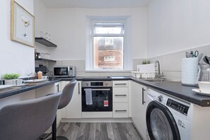 A compact kitchen with a microwave, oven, and dishwasher. There's a window above the sink, and a chair is positioned at the counter.