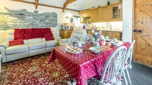 A cozy living room with a dining table set for a meal, a kitchen in the background, and a stone wall. - 2 Bed in Launceston (oc-congs) (Congdon's Shop)