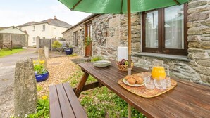 A stone building with a wooden table set for breakfast, including plates, glasses, and a basket of fruit. - 2 Bed in Launceston (oc-congs) (Congdon's Shop)