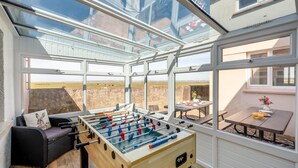 A room with a foosball table, a sofa with a cushion, and a table with a vase and drinks. - 5 Bed in Bideford (oc-codar) (Westward Ho!)