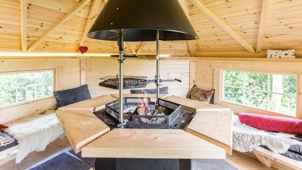 En mysig stuga med en central vedeldad kamin, träbjälkar i taket och ett fönster med utsikt över grönskan.