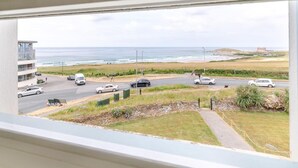 A coastal view from a window overlooking a road with cars, a beach, and a building with a balcony. - 3 Bed in Newquay (oc-headl) (Newquay)