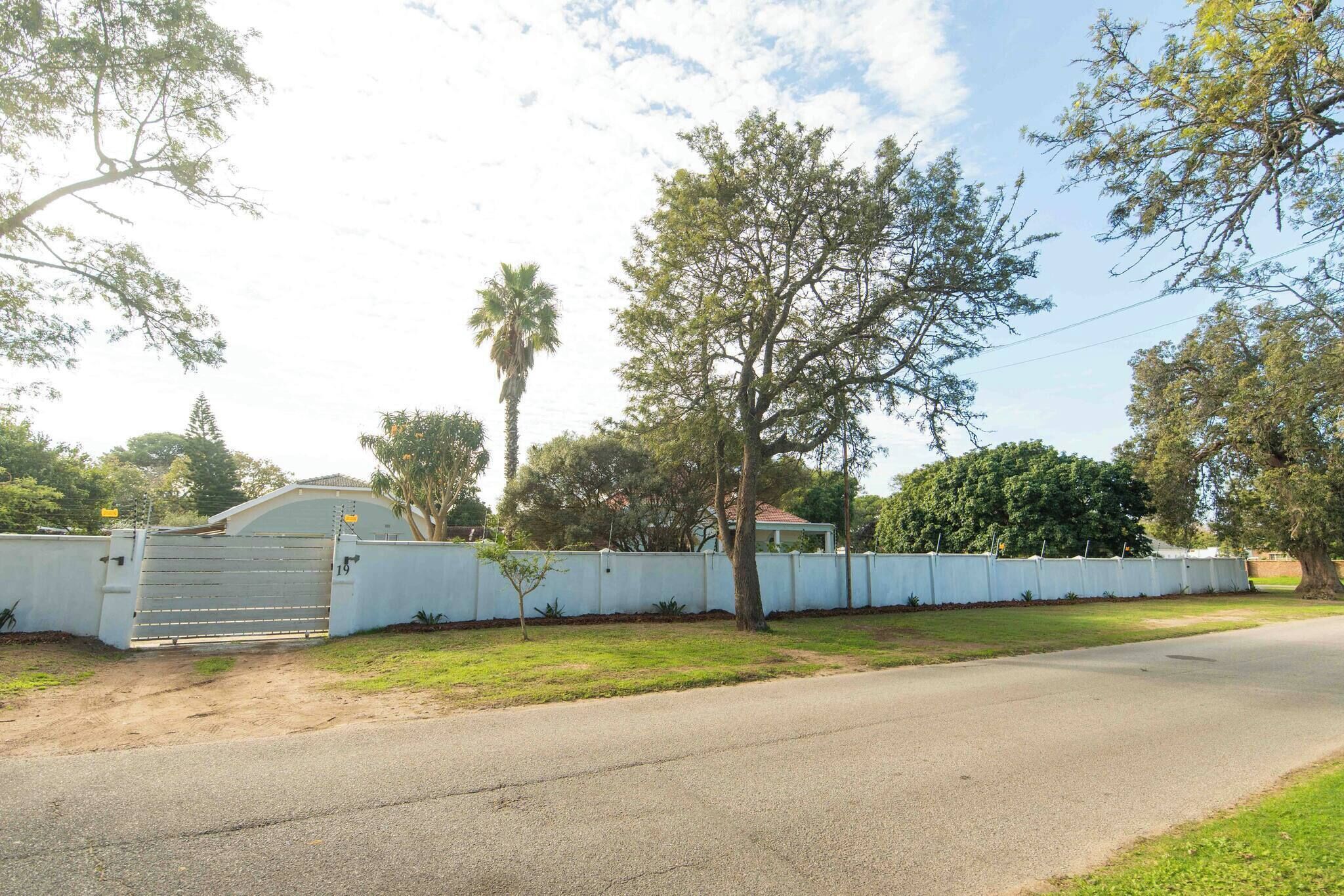 A gated residential area with a paved road, a white wall, and trees.