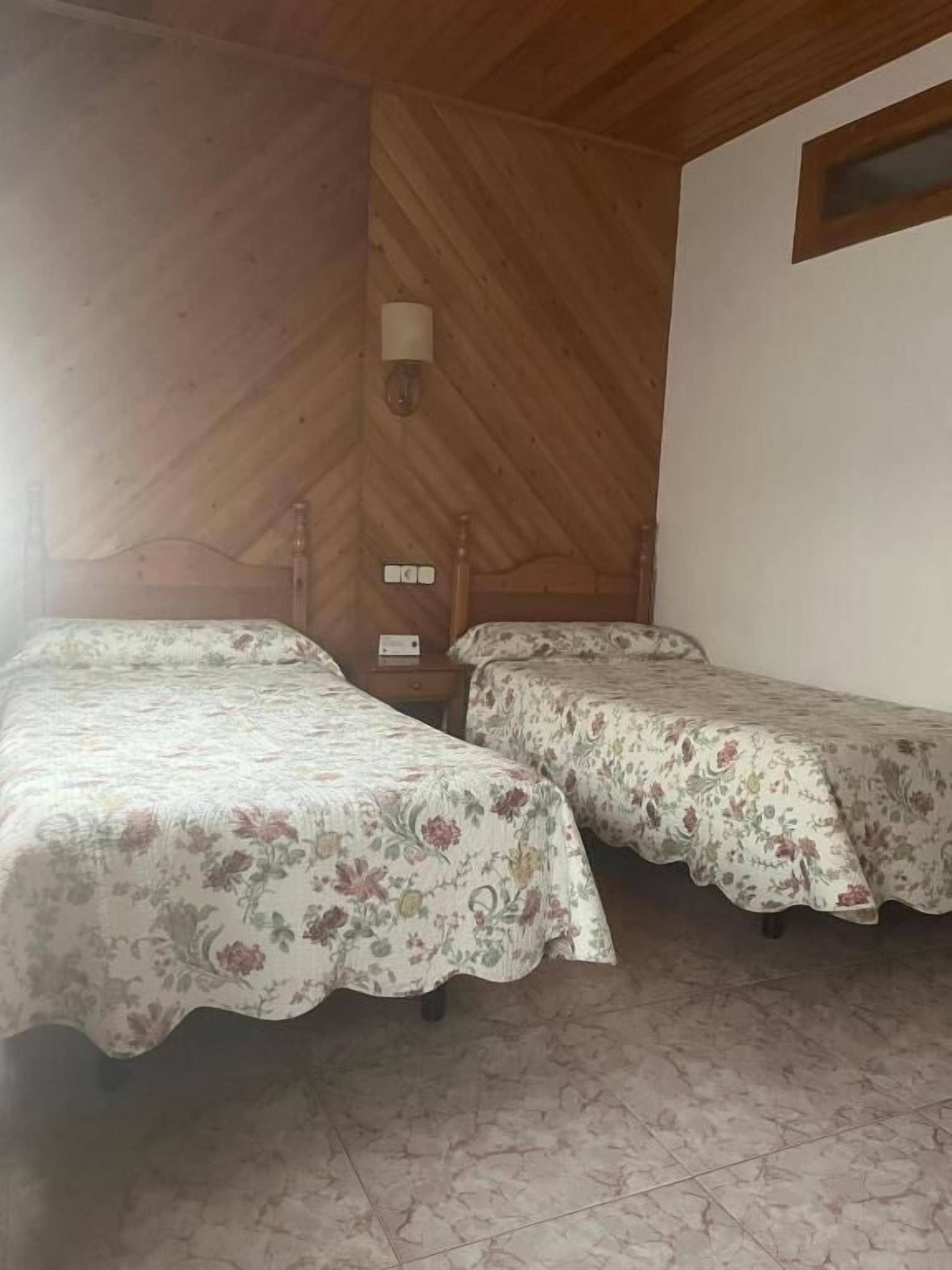 Una habitación con dos camas, paredes de madera, un ventanal pequeño y una lámpara fijada a la pared.