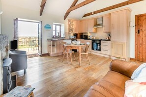 A spacious living area with a wooden floor, a dining table with chairs, a sofa, and a kitchen with wooden cabinets and a stove. - 1 Bed in Penzance (oc-henst) (Saint Buryan)