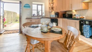 A kitchen with wooden cabinets, a dining table set with plates and cups, and a view of the outside. - 1 Bed in Penzance (oc-henst) (Saint Buryan)