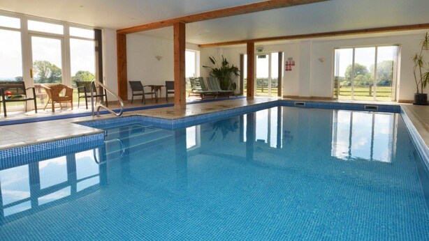 An indoor swimming pool with clear blue water, wooden beams, and a view of the outdoors. - 2 Bed in Sherborne (oc-folwo) (Folke)