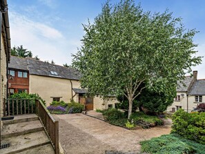 A courtyard with a large tree, a stone staircase, and a building with a sloped roof. - 2 Bed in Ilfracombe (oc-maltc) (West Down)