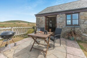 A stone building with a patio area featuring a wooden table, chairs, and a barbecue grill. - 1 Bed in Penzance (oc-ducks) (Saint Buryan)