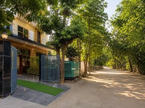 A tree-lined street with a modern building featuring a gated entrance and a green utility box. - Govind Residency (Greater Noida)