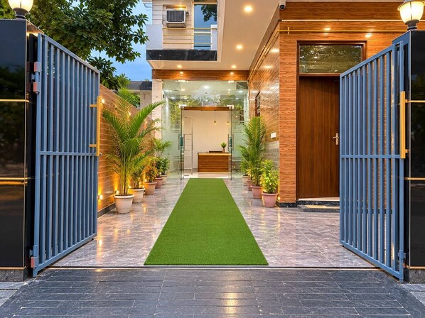 A modern building entrance with a reception desk, potted plants, and a green artificial turf path. - Govind Residency (Greater Noida)