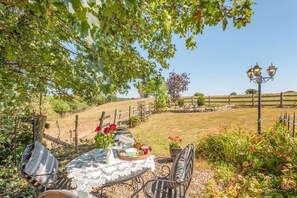 Uma área de refeições ao ar livre com uma mesa posta para dois, rodeada por vegetação e vista para um campo cercado.