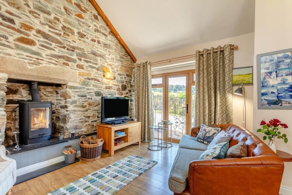 A cozy living room with a stone fireplace, a television on a wooden stand, a leather sofa, and a view through a glass door.