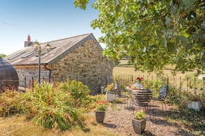 A stone building with a sloped roof, surrounded by a garden with various plants and a table with chairs.