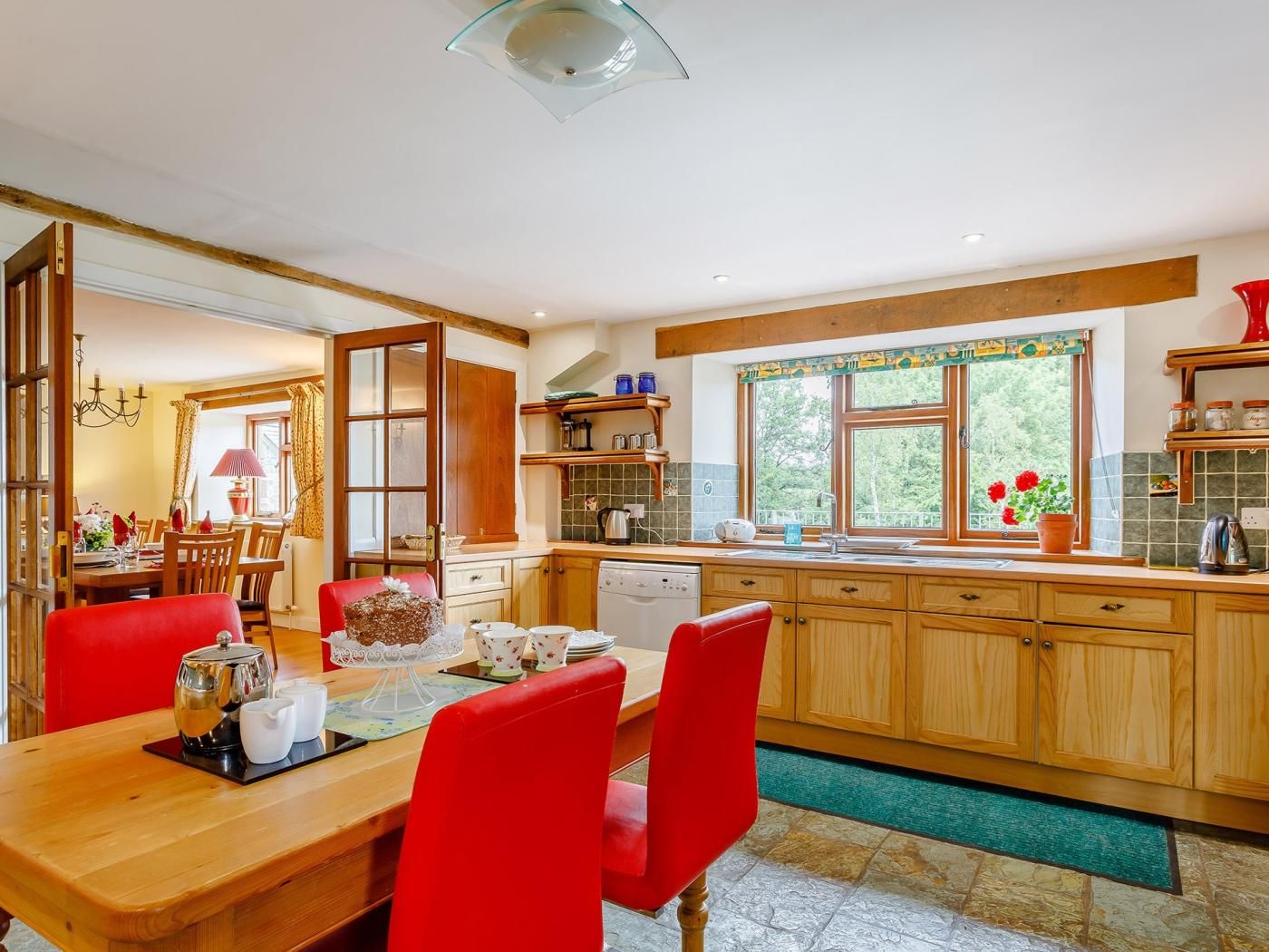 Una cocina con armarios de madera, una mesa de comedor con sillas rojas y un ventanal con vistas a zonas verdes.