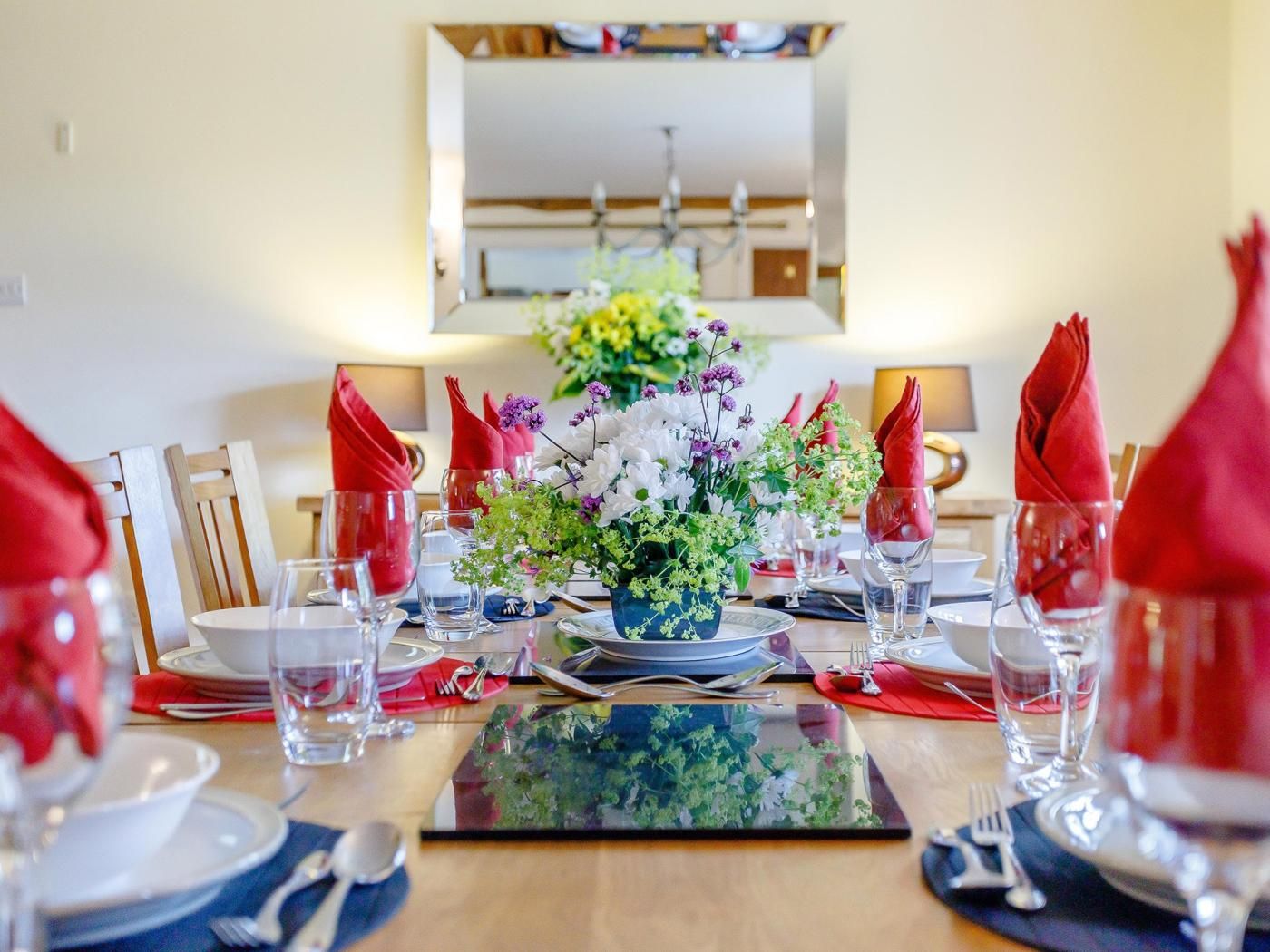 Een eettafel gedekt met rode servetten, helder glaswerk en een middelpunt van witte en paarse bloemen.