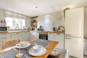 A modern kitchen with white cabinets, a dining table set with plates, glasses, and cutlery, and a clock on the wall. - 1 Bed in Beaworthy (oc-00782) (Sheepwash)