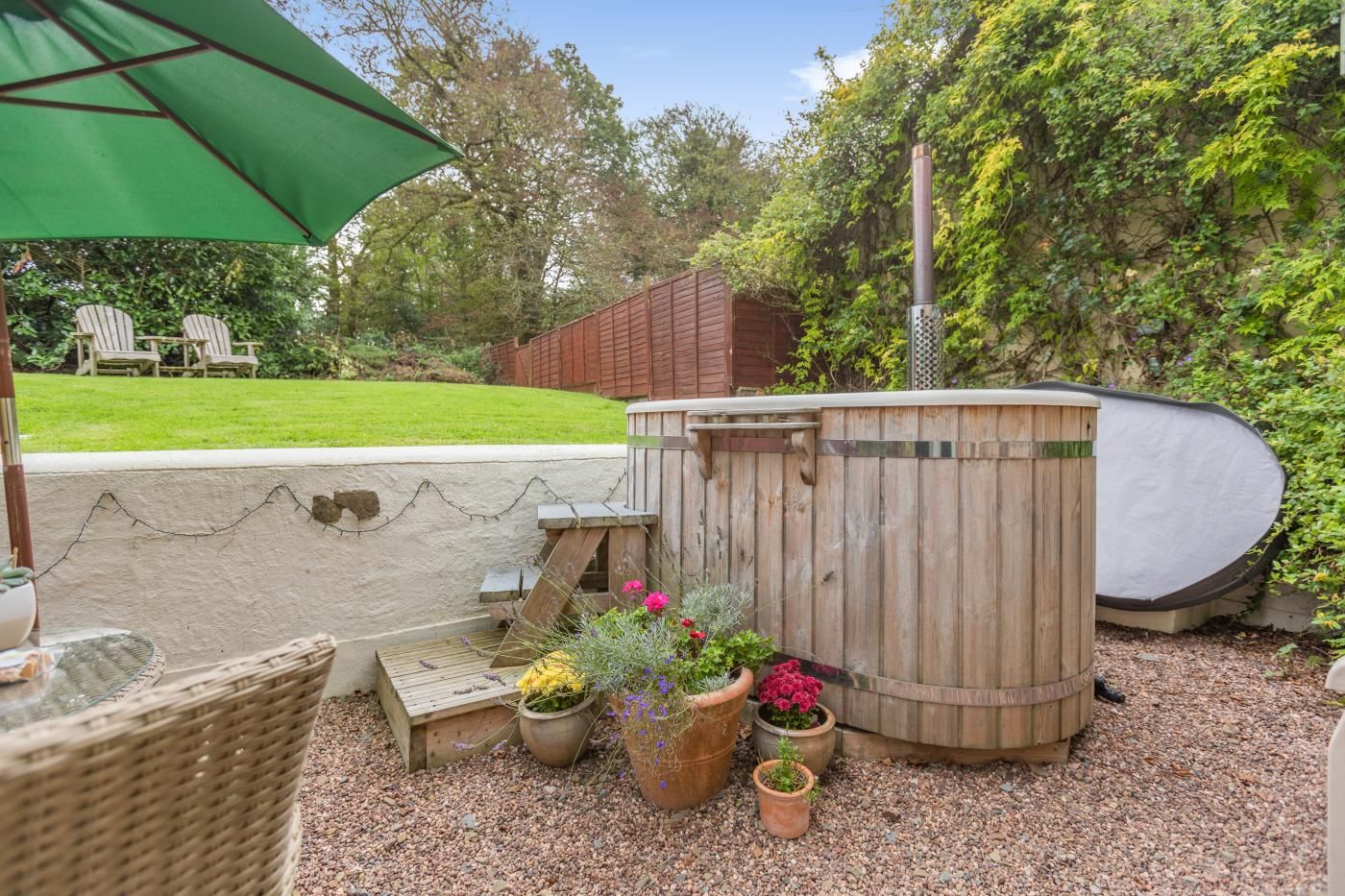 A wooden hot tub with a bench, surrounded by potted plants and a green umbrella.