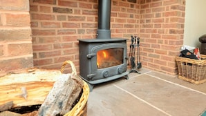 A wood-burning stove in a brick fireplace, a basket of firewood, and a wicker basket with items. - 3 Bed in Exeter (oc-harri) (Talaton)